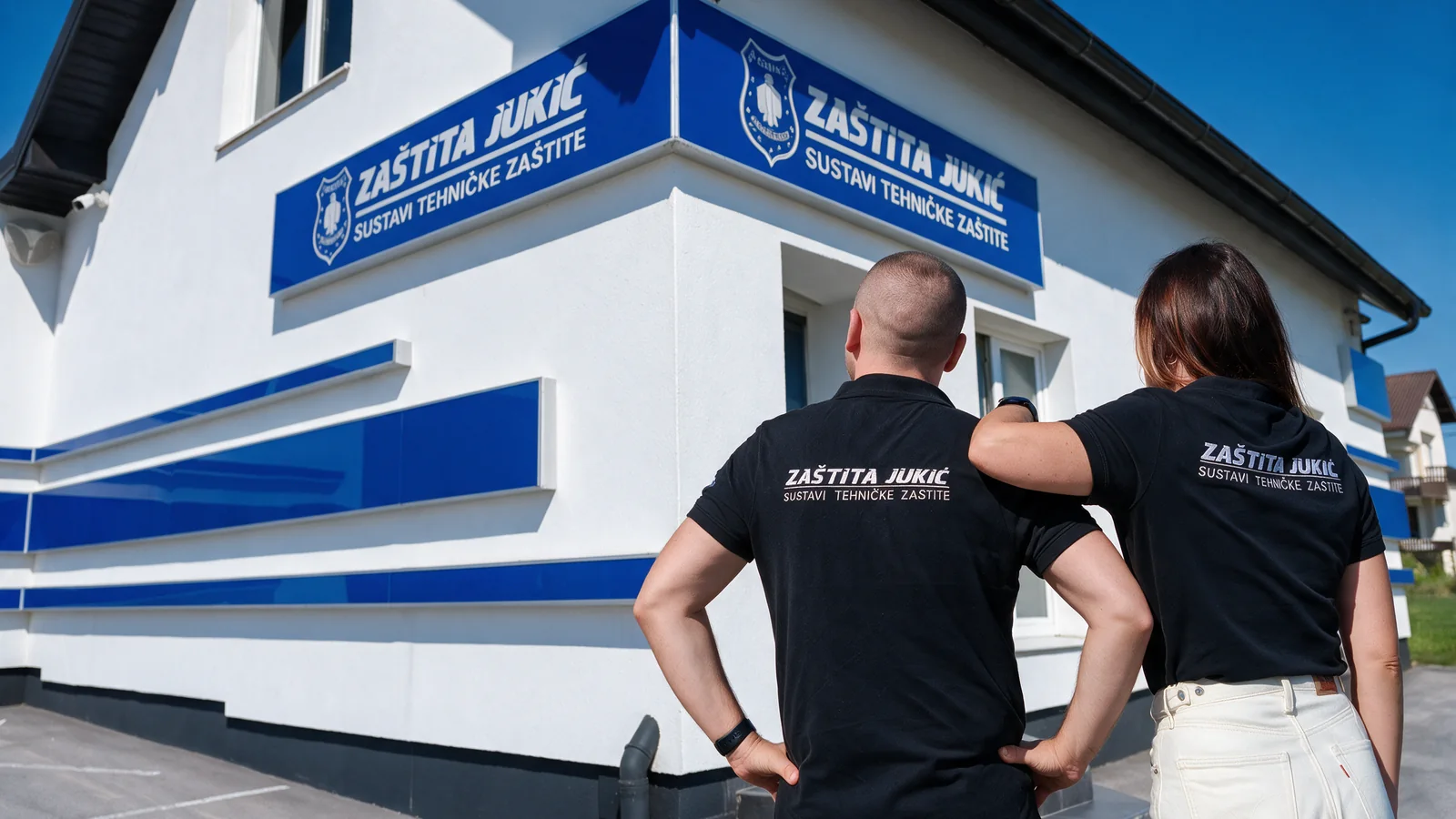 Dvoje djelatnika Zaštite Jukić u brendiranim majicama pred sjedištem tvrtke s natpisom Zaštita Jukić – sustavi tehničke zaštite